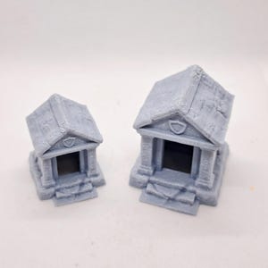 May include: Two gray miniature temple models with detailed architectural features. Each model has a pitched roof, columns, and a recessed entrance. The temples are likely for tabletop gaming or model building.
