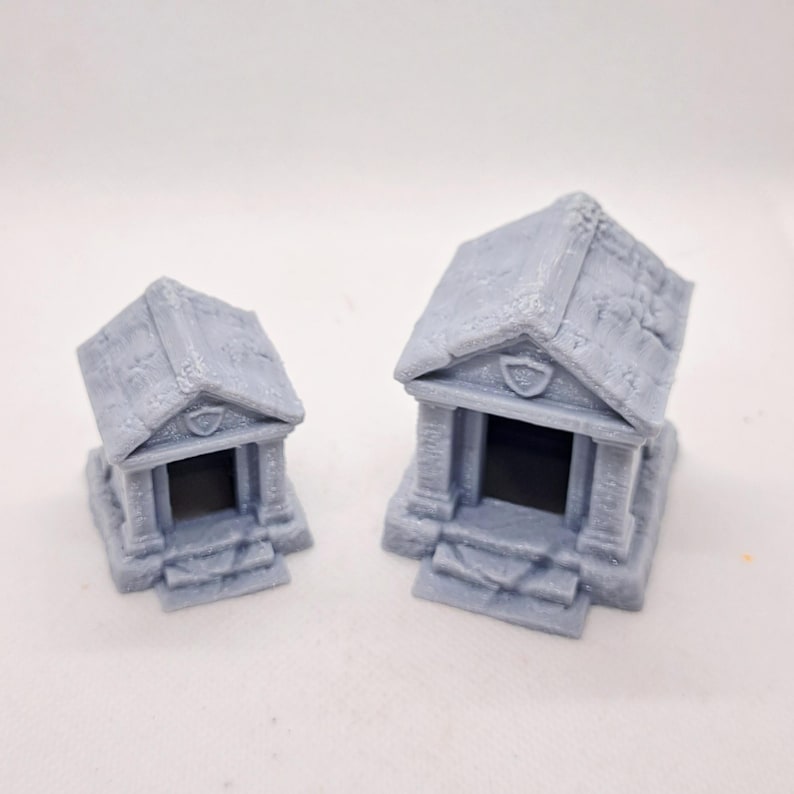 May include: Two gray miniature temple models with detailed architectural features. Each model has a pitched roof, columns, and a recessed entrance. The temples are likely for tabletop gaming or model building.