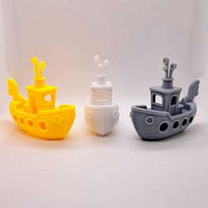 May include: Three toy boats in yellow, white, and gray. Each boat has a cabin, smokestacks, and portholes. The yellow boat has a large fin on the side. The boats are small and appear to be 3D printed.