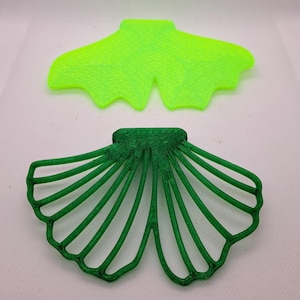 May include: Two leaf-shaped objects. One is a solid, bright green color. The other is a darker green, open-work design with radiating lines, resembling a fan or leaf. Both are on a white surface.