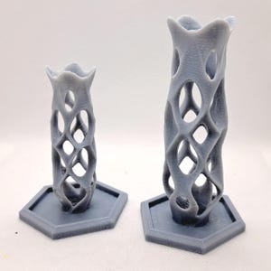 May include: Two gray, 3D-printed decorative vases. The vases have an open, lattice-like design with a flared top and sit on hexagonal bases. One vase is taller than the other. The vases are made of a matte gray material.