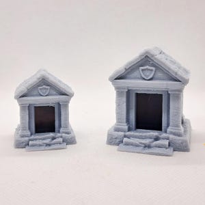 May include: Two gray miniature mausoleums with detailed architectural features. Each has a pitched roof, columns, and a shield emblem above the entrance. The mausoleums are designed with steps leading up to the dark rectangular openings.