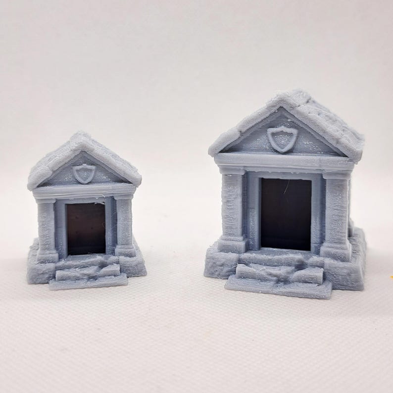May include: Two gray miniature mausoleums with detailed architectural features. Each has a pitched roof, columns, and a shield emblem above the entrance. The mausoleums are designed with steps leading up to the dark rectangular openings.