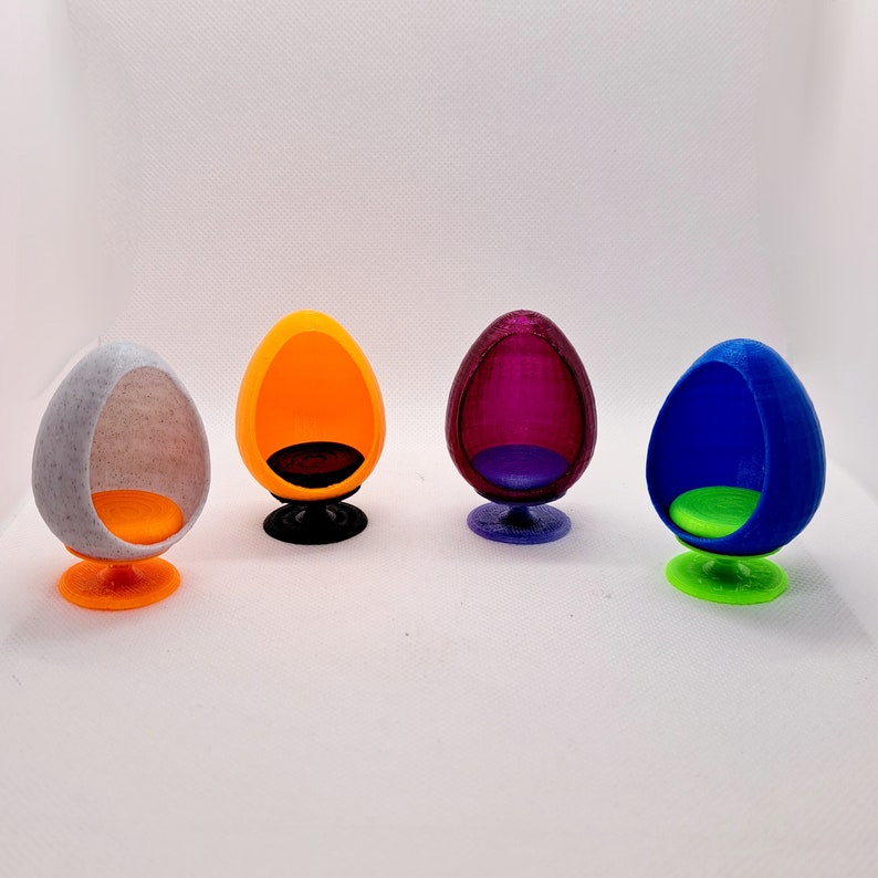 May include: Four miniature egg-shaped chairs in different colours: white, orange, purple, and blue. Each chair has a round base and a smooth, rounded design.