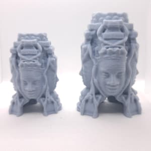 May include: Two gray, 3D-printed sculptures of a multi-faced figure with a serene expression. The sculptures have a detailed, architectural design with multiple faces and a crown-like structure. The larger sculpture is approximately 5 inches tall.