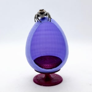 May include: A miniature purple egg-shaped chair with a dark purple seat and a small spider on top.