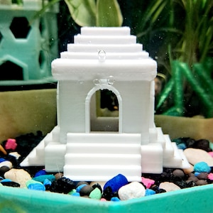 May include: White plastic miniature temple with a small opening in the front, sitting on a bed of colorful pebbles.