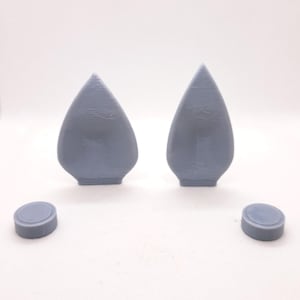 May include: Two gray, teardrop-shaped objects and two small, round objects are displayed against a white background. The teardrop shapes have a textured surface and a flat base. The round objects are small, flat discs.