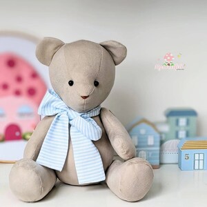 May include: A beige plush teddy bear wearing a blue and white striped scarf. The bear is sitting on a white surface with a pink and white mushroom-shaped object in the background.