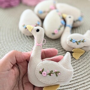 May include: A felt goose ornament with embroidered pink flowers and green leaves. The goose is light beige with a yellow beak and feet.