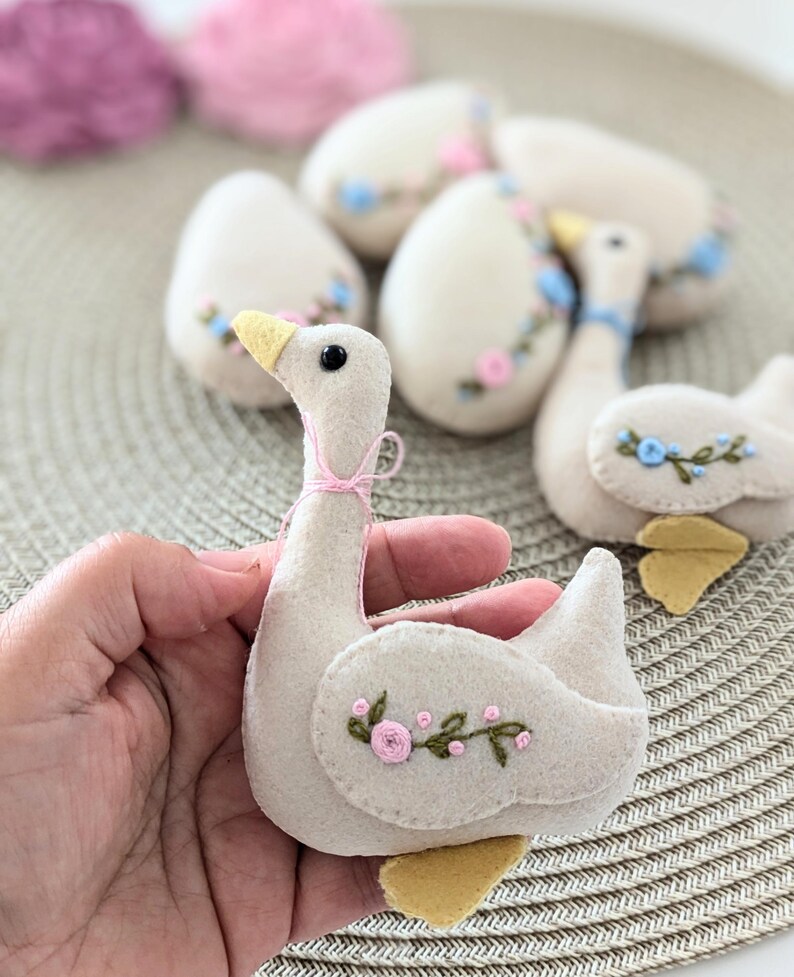 May include: A felt goose ornament with embroidered pink flowers and green leaves. The goose is light beige with a yellow beak and feet.