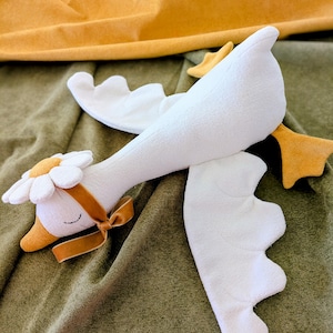 May include: A soft white goose toy with a daisy flower on its head and a brown ribbon tied around its neck. The goose has yellow feet and a yellow beak. The toy is on a green surface with yellow fabric in the background.