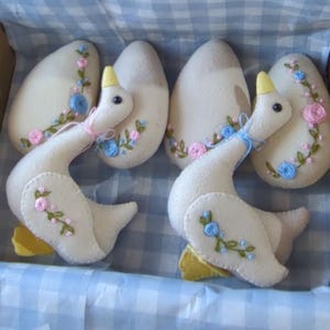 May include: Three white felt goose ornaments with embroidered flowers and leaves in pink, blue, and green. The geese have yellow beaks and feet.