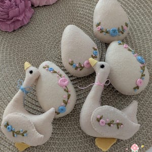 May include: Five felt Easter eggs and two felt geese, all in a light beige colour with embroidered floral designs in pink and blue. The eggs and geese are arranged on a woven brown background.