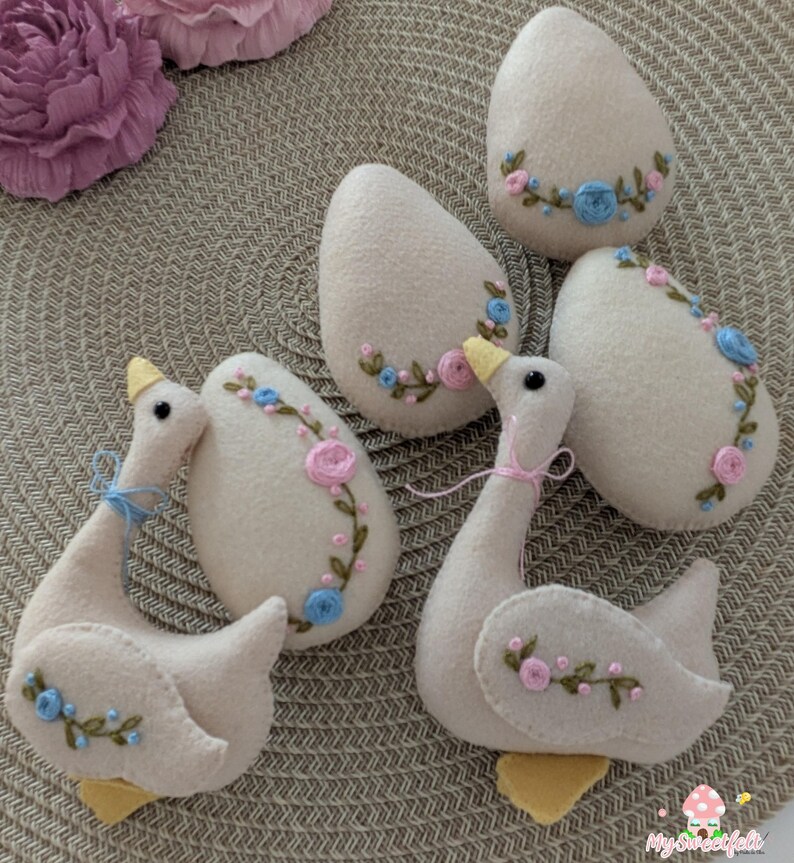 May include: Five felt Easter eggs and two felt geese, all in a light beige colour with embroidered floral designs in pink and blue. The eggs and geese are arranged on a woven brown background.