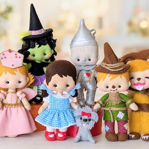 Könnte beinhalten: Eine Sammlung von sieben Filzpuppen, die Charaktere aus dem Zauberer von Oz darstellen, darunter eine Fee in Rosa, eine grüne Hexe mit schwarzem Hut, ein Mädchen im blau karierten Kleid, ein silberner Roboter, ein kleiner grauer Hund, eine Vogelscheuche und ein Löwe.