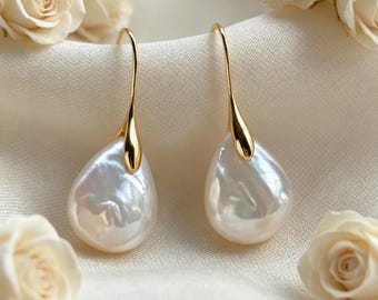 May include: A pair of drop earrings featuring teardrop-shaped white pearls suspended from polished gold-tone metal hooks. The pearls have a lustrous, iridescent surface and are securely attached to the curved gold metal findings.