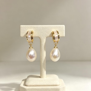 May include: A pair of gold hoop earrings featuring a white inlay and a dangling teardrop-shaped pearl. The hoops have a polished gold finish with a smooth white center band, and the pearls exhibit a lustrous, iridescent white surface.