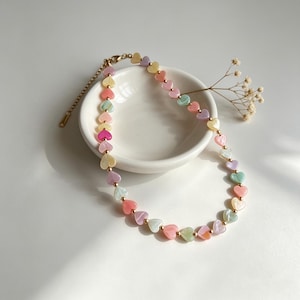 May include: A pastel-coloured beaded necklace with heart-shaped beads in shades of pink, yellow, green, and purple. The necklace has a gold-coloured chain and clasp, and is displayed on a white surface.