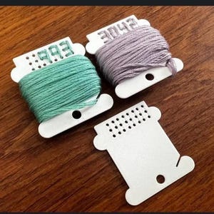 Puede incluir: Tres carretes de hilo de bordar en tonos verdes y morados, con bobinas blancas. Una bobina está vacía. Los carretes están etiquetados con números. El fondo es una superficie de madera.