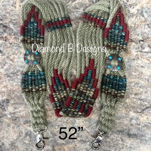 May include: A 52-inch long woven strap featuring sage green yarn with geometric patterns in maroon, teal, and brown. The strap includes two silver-toned circular conchos with turquoise accents and metal swivel lobster clasps at each end. Text reads Diamond B Designs.