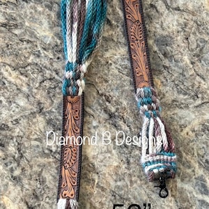 May include: A 52-inch strap featuring braided yarn in teal, brown, and white, connected to two sections of brown leather with floral tooling. The strap includes a silver-toned metal ring and two black swivel clips. Text reads Diamond B Designs.