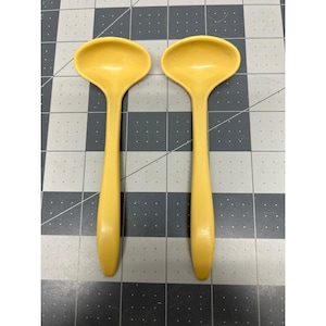 May include: Two matching yellow ladles with smooth, rounded bowls and long, slender handles. These kitchen utensils feature a solid, uniform color and a simple, functional design with a curved shape at the top of the handle for a comfortable grip.