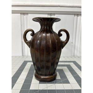 May include: A bronze-colored vase with a ribbed design and two curved handles. The vase has a wide, flared top and a rounded base. The overall shape is symmetrical and elegant, suitable for home decor.