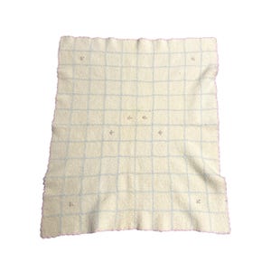 May include: Cream-colored, square blanket with a grid pattern of light blue lines. The blanket has a pink, scalloped edge and small, embroidered floral accents at the grid intersections. The fabric appears soft and textured.