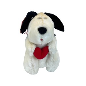May include: A plush dog toy with a cream-colored body, black ears, and a black nose. The dog is holding a red heart. The toy has a small red tag attached to its ear. The dog's eyes are closed, and it has a small red mouth.