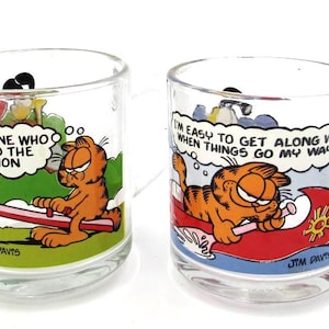 Puede incluir: Dos tazas de cristal transparente con el personaje de dibujos animados Garfield. La taza izquierda muestra a Garfield en un balancín con el texto I'M NOT ONE WHO RISES TO THE OCCASION. La derecha muestra a Garfield en una canoa roja con el texto I'M EASY TO GET ALONG WITH WHEN THINGS GO MY WAY.