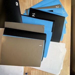 May include: A collection of various colored sheets of paper, including black, blue, tan, and white. Some sheets have numbers printed on them, such as 1, 2, 5, and 14. The papers are arranged on a wooden surface.