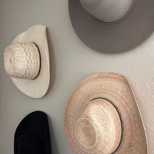 May include: Four cowboy hats of varying styles and colors are mounted on a light gray wall. The hats include a black felt hat, a light gray felt hat, and two straw hats, one with a decorative woven pattern.
