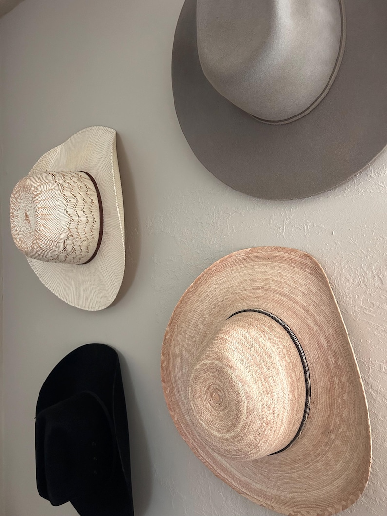May include: Four cowboy hats of varying styles and colors are mounted on a light gray wall. The hats include a black felt hat, a light gray felt hat, and two straw hats, one with a decorative woven pattern.