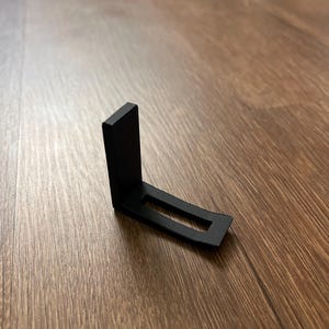 May include: A black metal bracket with an L-shape design. The bracket has a rectangular base with a rectangular cutout. The vertical piece is also rectangular. The bracket is on a wooden surface.
