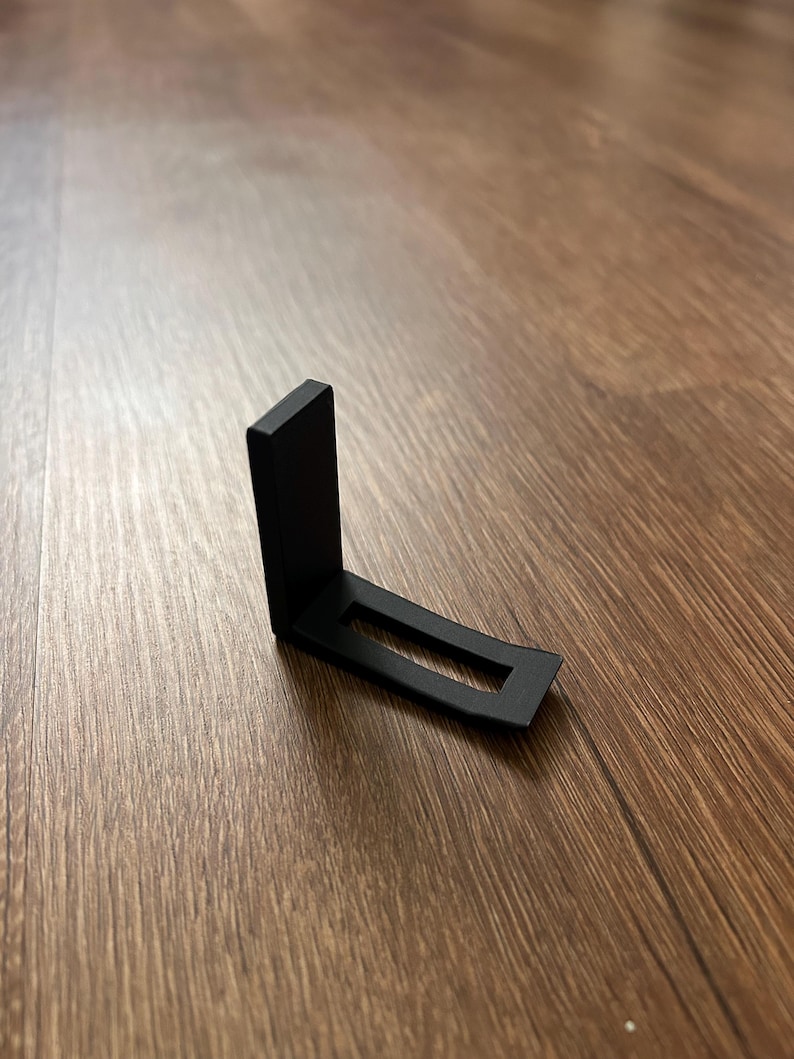 May include: A black metal bracket with an L-shape design. The bracket has a rectangular base with a rectangular cutout. The vertical piece is also rectangular. The bracket is on a wooden surface.