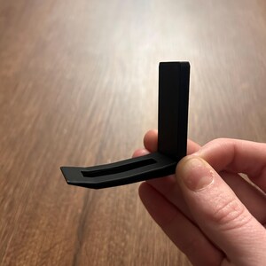 May include: A black, L-shaped bracket with a curved base and a vertical support. The bracket is held against a wooden surface. The design is simple and modern, with clean lines and a matte finish.
