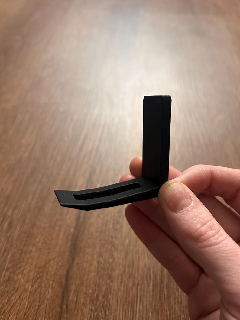 May include: A black, L-shaped bracket with a curved base and a vertical support. The bracket is held against a wooden surface. The design is simple and modern, with clean lines and a matte finish.