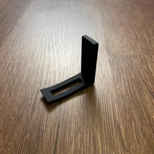 May include: A black, right-angled bracket with a rectangular cutout. The bracket is made of a matte material and sits on a wood-grain surface. The design is simple and functional, likely for support or mounting.