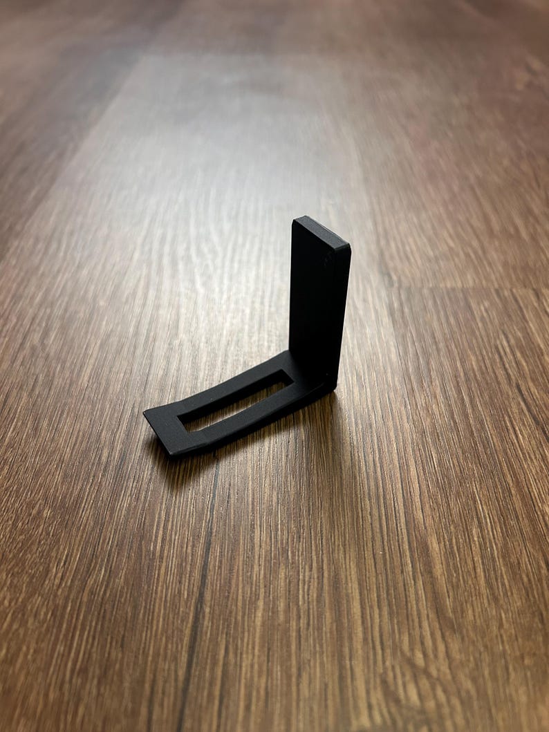 May include: A black, right-angled bracket with a rectangular cutout. The bracket is made of a matte material and sits on a wood-grain surface. The design is simple and functional, likely for support or mounting.