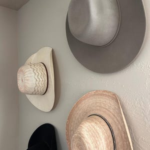 Könnte beinhalten: Vier Cowboyhüte in verschiedenen Stilen und Farben sind an einer Wand befestigt. Die Hüte umfassen einen hellgrauen Filzhut, einen Strohhut mit schwarzem Band, einen weißen Hut mit gemustertem Oberteil und einen schwarzen Hut.