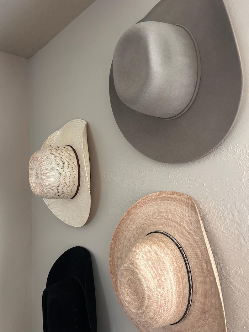 May include: Four cowboy hats of different styles and colors are mounted on a wall. The hats include a light gray felt hat, a straw hat with a black band, a white hat with a patterned crown, and a black hat.
