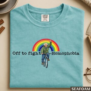 May include: Seafoam green crew neck t-shirt. The graphic shows a person on a bicycle under a rainbow, with the text "Off to fight Homophobia". The t-shirt is a Comfort Colors brand.