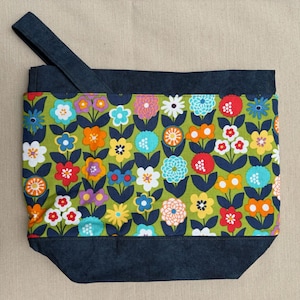 May include: A tote bag with a colorful floral pattern on a green background. The flowers are in various colors, including red, orange, yellow, and blue. The top and bottom of the bag are a dark blue fabric, and it has a matching strap.