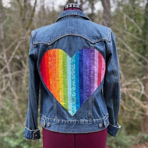 May include: A denim jacket with a large, colorful, rainbow-striped heart design on the back. The heart is made of various patterned fabrics in shades of red, orange, yellow, green, blue, and purple. The jacket has a classic collar and button closures.
