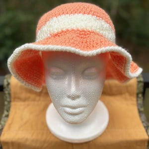 May include: A handmade crochet sun hat in peach and white. The hat features a wide, floppy brim and a striped design around the crown. The hat is displayed on a white foam head, showcasing its shape and style. The hat is a summer accessory.