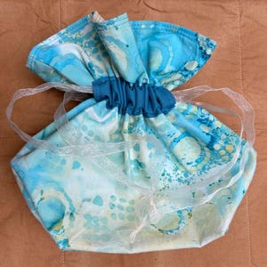 May include: A handmade drawstring bag with a blue and white watercolor pattern. The bag is gathered at the top with a dark blue band and has a sheer white ribbon drawstring. The fabric has a marbled effect.