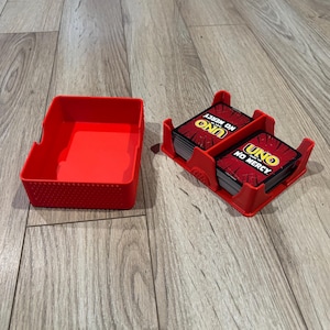 May include: A red plastic card holder with a textured exterior and a matching red plastic tray. The card holder contains two stacks of Uno No Mercy playing cards, each featuring the game logo and text on the top card of the deck.