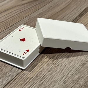 May include: A deck of playing cards housed in a white plastic case with the lid partially removed. The top card is an ace of hearts, featuring a white background with subtle grey speckles and red heart symbols and the letter A in the corners.