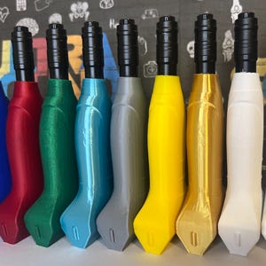 May include: A row of nine vertical tattoo machine grips in various colors including blue, red, green, teal, gray, yellow, gold, and white. Each grip features a black cylindrical top section and a smooth, contoured handle with a flat base.